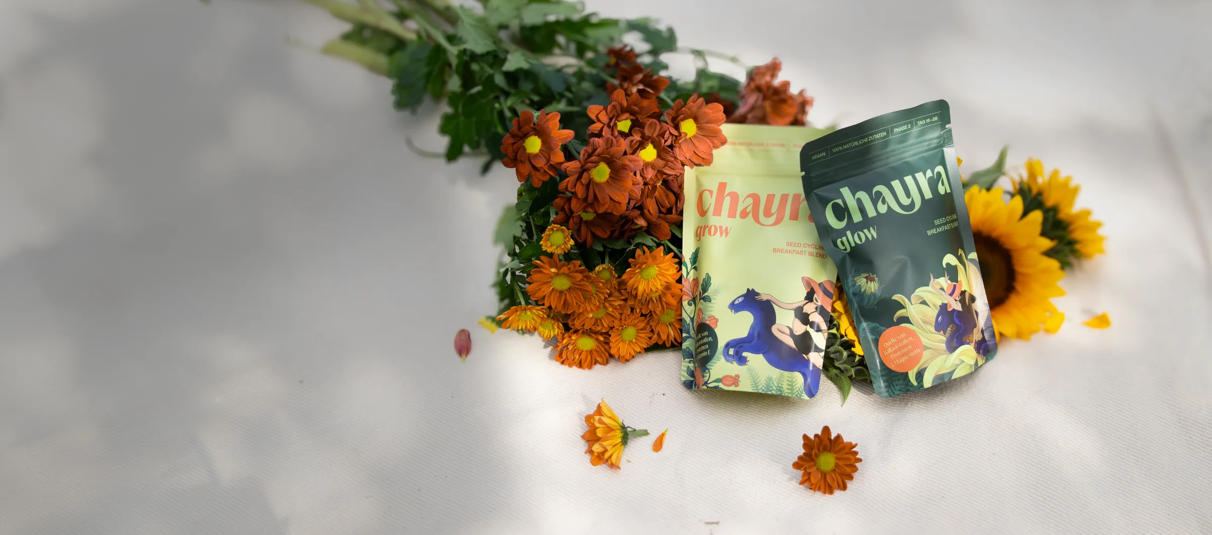 CHAYRA GROW und GLOW auf einer hellen Decke, daneben ein Strauss mit orangen Blumen und Sonnenblumen